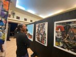 Seorang pria berambut gondrong berbaju hitam sedang berdiri dan mengamati lukisan bergaya kolase pada panel hitam di pameran seni di Hotel Srikandi Pacitan.
