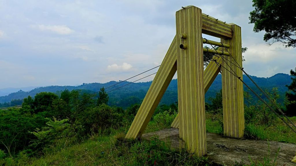 Struktur kayu wahana flying fox yang usang di Bukit Cakrawala Pacitan, dikelilingi rumput liar dengan latar belakang perbukitan hijau dan langit mendung.