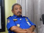 Kabid Damkar Satpol PP Pacitan, Sugito, yang mengenakan seragam biru lengkap dengan atribut dan papan nama, sedang duduk bersedekap sambil berbicara di depan mikrofon dalam sebuah sesi wawancara berlatar belakang tirai abu-abu.