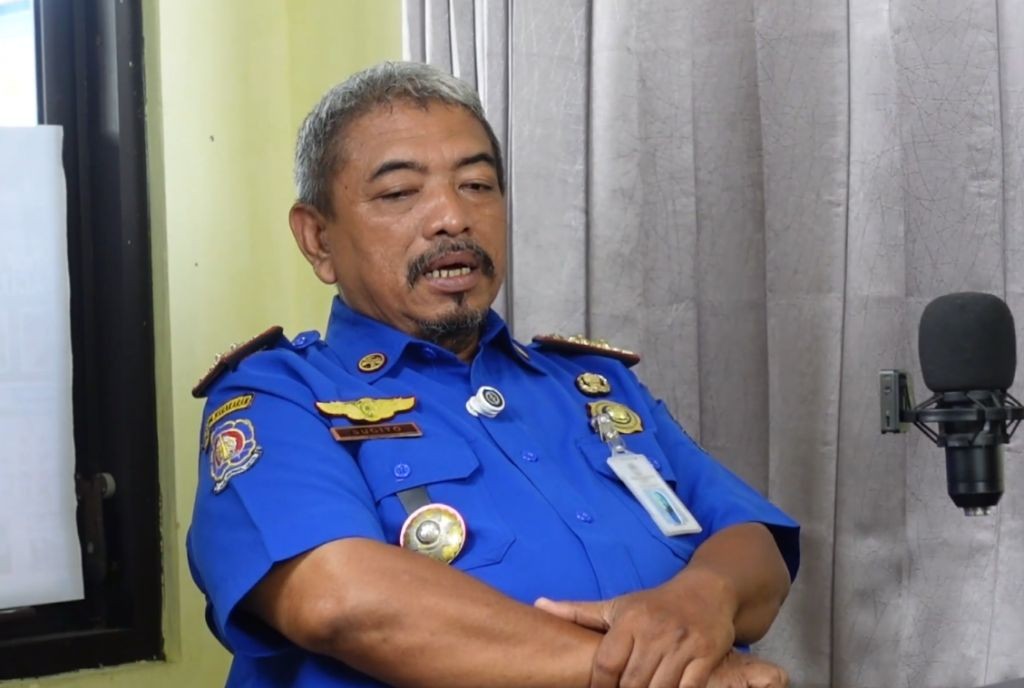 Kabid Damkar Satpol PP Pacitan, Sugito, yang mengenakan seragam biru lengkap dengan atribut dan papan nama, sedang duduk bersedekap sambil berbicara di depan mikrofon dalam sebuah sesi wawancara berlatar belakang tirai abu-abu.