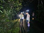 Sejumlah petugas kepolisian berseragam lengkap sedang melakukan olah Tempat Kejadian Perkara (TKP) kecelakaan lalu lintas pada malam hari di jalan raya beraspal yang sempit dan dikelilingi pepohonan rimbun di Pacitan. Seorang petugas tampak memegang alat ukur roda berjalan.