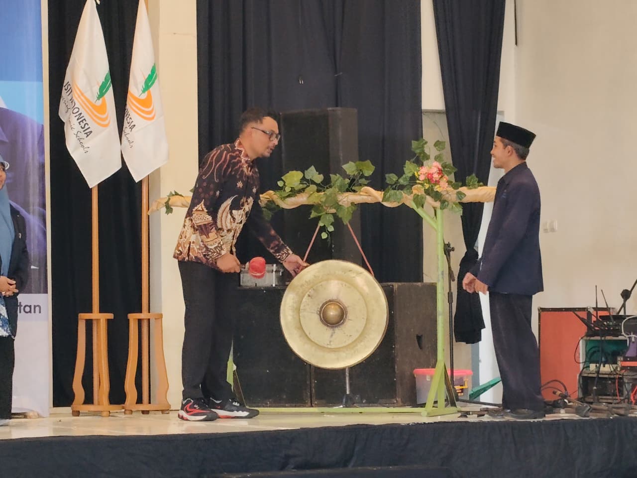 Kepala Dinas Pendidikan Kabupaten Pacitan, Khemal Pandu Pratikna, yang mengenakan kemeja batik, sedang menabuh gong besar di atas panggung sebagai simbol pembukaan acara Musyawarah Daerah ke-3 Jaringan Sekolah Islam Terpadu (JSIT) Pacitan, disaksikan oleh seorang pria berpeci di sampingnya.