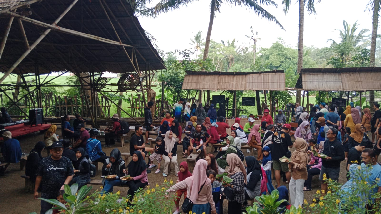 Pemandangan luas suasana Pasar Beling yang ramai dipadati pengunjung. Terlihat struktur bangunan bambu besar di sebelah kiri dan deretan gubuk bambu penjual di sebelah kanan, dengan banyak orang duduk santai dan berinteraksi di area terbuka yang dikelilingi pohon kelapa.