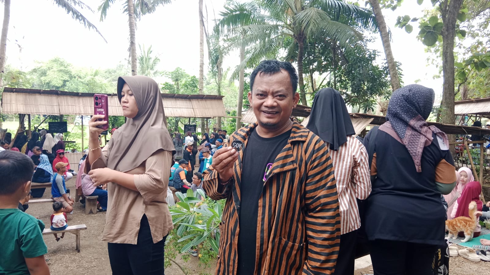 Cristian Ade Candra, seorang pria tersenyum mengenakan baju lurik bergaris cokelat hitam, memegang dan menunjukkan sebuah koin atau token bertuliskan "50K". Ia berdiri dengan latar belakang suasana Pasar Beling yang ramai pengunjung.