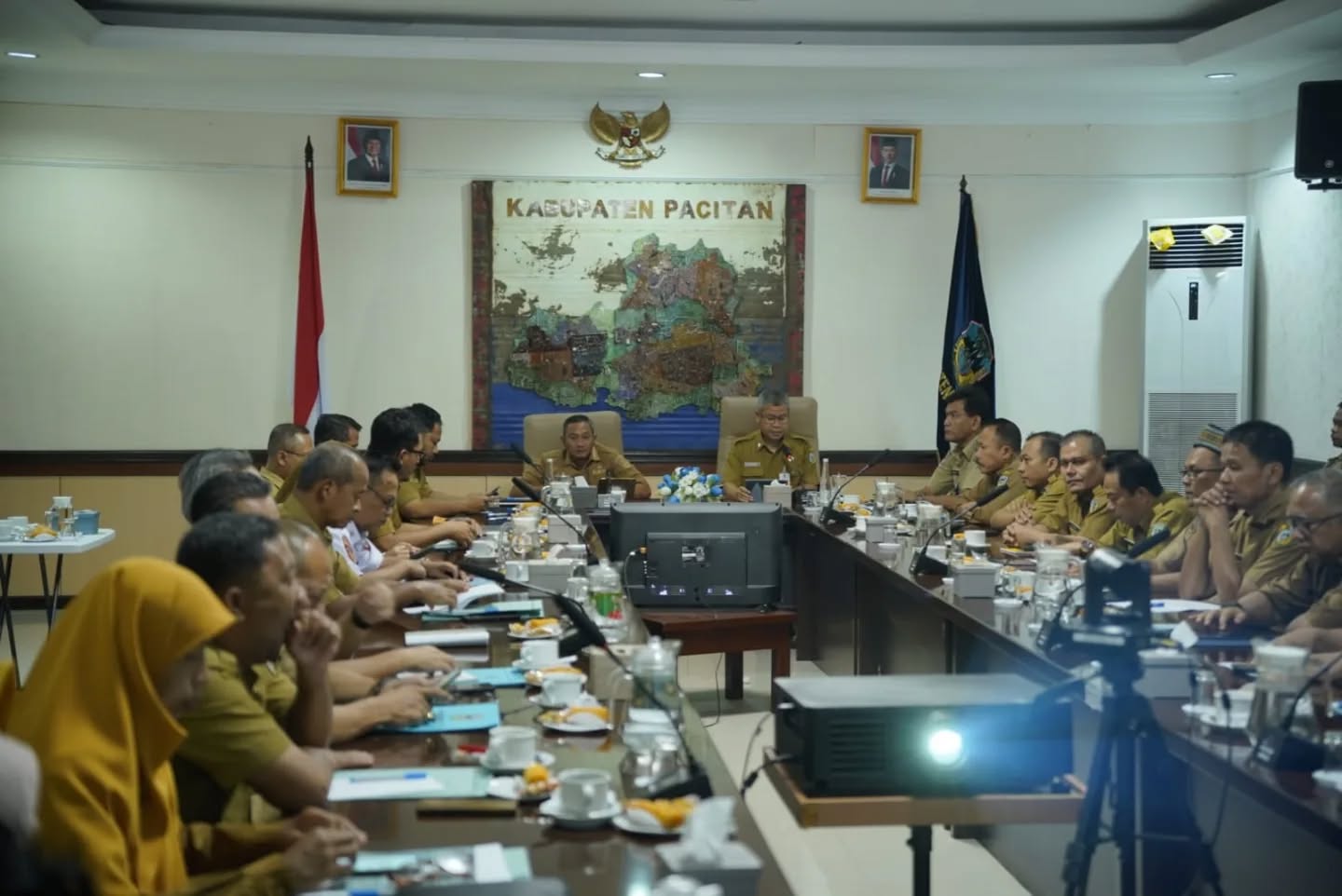 Suasana rapat kerja di ruangan pertemuan pemerintah daerah, di mana Bupati Pacitan, Indrata Nur Bayuaji (duduk di tengah ujung meja), memimpin jalannya diskusi. Puluhan pria berseragam dinas khaki (ASN) duduk mengelilingi meja konferensi panjang yang dilengkapi mikrofon dan dokumen. Di dinding belakang Bupati terdapat peta besar bertuliskan "KABUPATEN PACITAN" yang diapit oleh foto resmi Presiden dan Wakil Presiden Republik Indonesia. Sebuah proyektor terlihat di bagian depan meja.