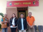 Empat orang—tiga pria dan satu wanita—berdiri berjejer di depan pintu masuk bangunan kantor polisi yang di atasnya terdapat papan merah bertuliskan "UNIT I / PIDUM" dan lambang kepolisian Jawa Timur. Pria paling kiri mengenakan rompi cokelat bertuliskan "ADVOCAT", wanita di sebelahnya berjaket hitam, pria di tengah berjas hitam, dan pria paling kanan yang tampak lebih tua mengenakan kaos polo oranye.