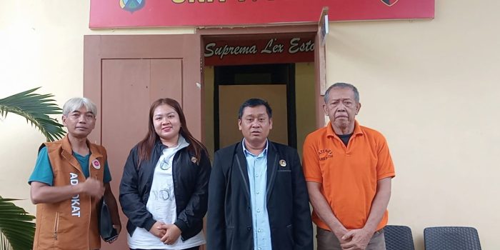 Empat orang—tiga pria dan satu wanita—berdiri berjejer di depan pintu masuk bangunan kantor polisi yang di atasnya terdapat papan merah bertuliskan "UNIT I / PIDUM" dan lambang kepolisian Jawa Timur. Pria paling kiri mengenakan rompi cokelat bertuliskan "ADVOCAT", wanita di sebelahnya berjaket hitam, pria di tengah berjas hitam, dan pria paling kanan yang tampak lebih tua mengenakan kaos polo oranye.