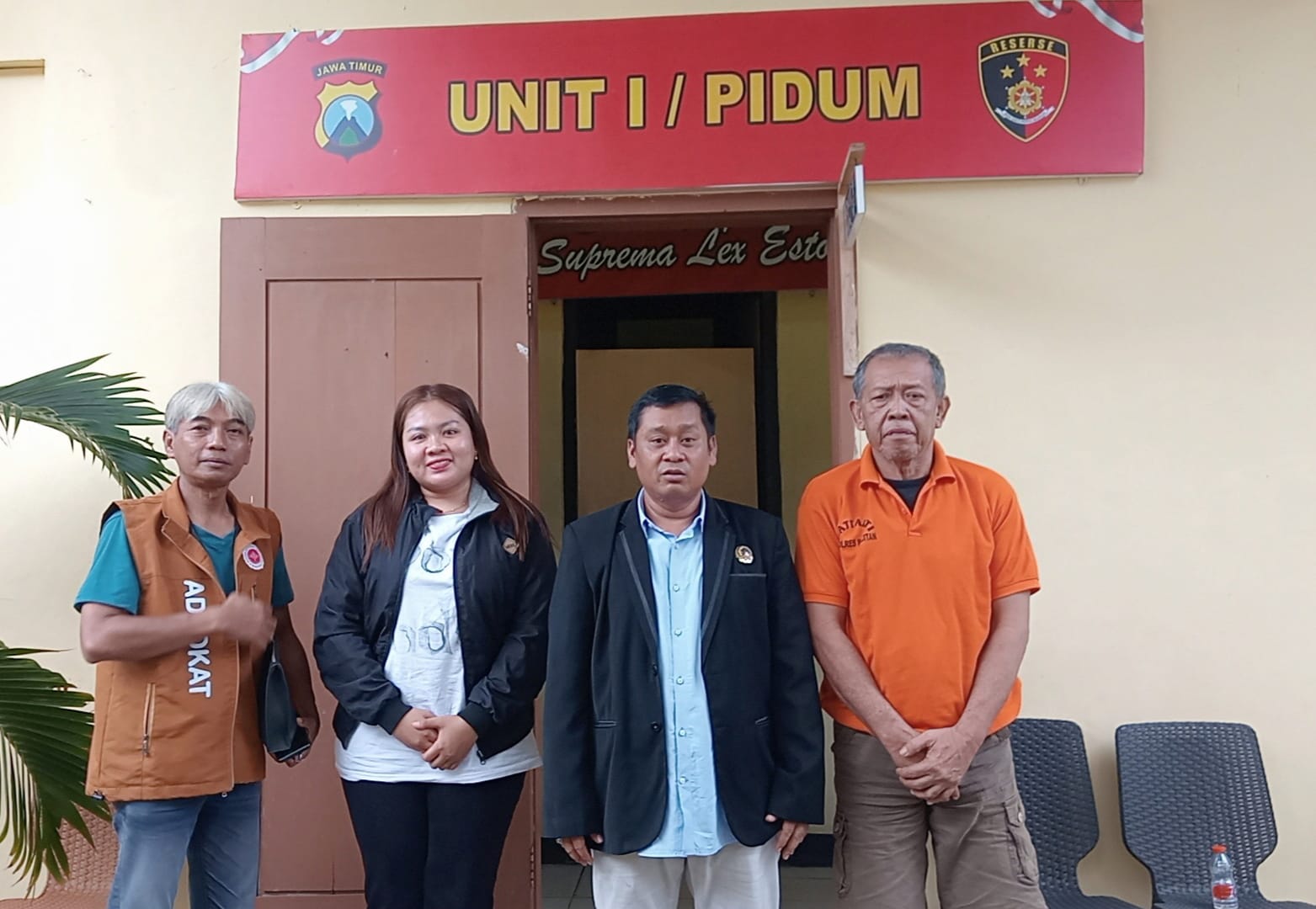 Empat orang—tiga pria dan satu wanita—berdiri berjejer di depan pintu masuk bangunan kantor polisi yang di atasnya terdapat papan merah bertuliskan "UNIT I / PIDUM" dan lambang kepolisian Jawa Timur. Pria paling kiri mengenakan rompi cokelat bertuliskan "ADVOCAT", wanita di sebelahnya berjaket hitam, pria di tengah berjas hitam, dan pria paling kanan yang tampak lebih tua mengenakan kaos polo oranye.