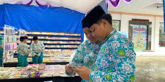 Dua pelajar laki-laki berseragam batik hijau bermotif dan berpeci hitam sedang fokus mengamati sebuah buku di stan bazar buku yang dipenuhi tumpukan buku di bawah tenda beratapkan kain biru dan berhias kain ungu.