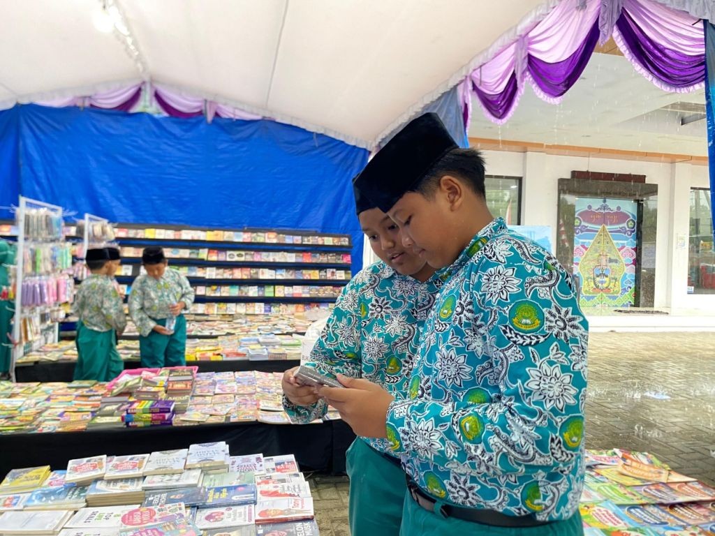 Dua pelajar laki-laki berseragam batik hijau bermotif dan berpeci hitam sedang fokus mengamati sebuah buku di stan bazar buku yang dipenuhi tumpukan buku di bawah tenda beratapkan kain biru dan berhias kain ungu.