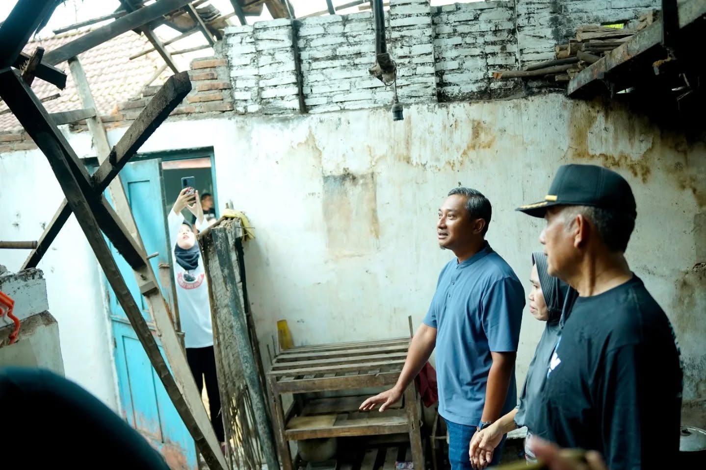 Bupati Pacitan Indrata Nur Bayuaji mengenakan kemeja biru menunjuk ke arah puing bangunan yang runtuh saat meninjau lokasi terdampak gempa bumi di dalam sebuah rumah warga yang rusak berat.
