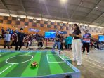 Suasana pertandingan robot soccer di dalam gedung olahraga. Seorang siswi berbaju hitam dan celana putih sedang mengendalikan robot menggunakan ponsel pintarnya di arena mini, disaksikan oleh peserta lain dan panitia.
