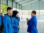 Ketua STKIP PGRI Pacitan, Bakti Sutopo, berjas biru, memberikan salam dengan menangkupkan tangan di depan dada kepada dua perwakilan mahasiswa KKN berjas biru dalam acara pelepasan di dalam gedung aula yang luas. Sejumlah mahasiswa lain berdiri berbaris di latar belakang menyaksikan prosesi tersebut.