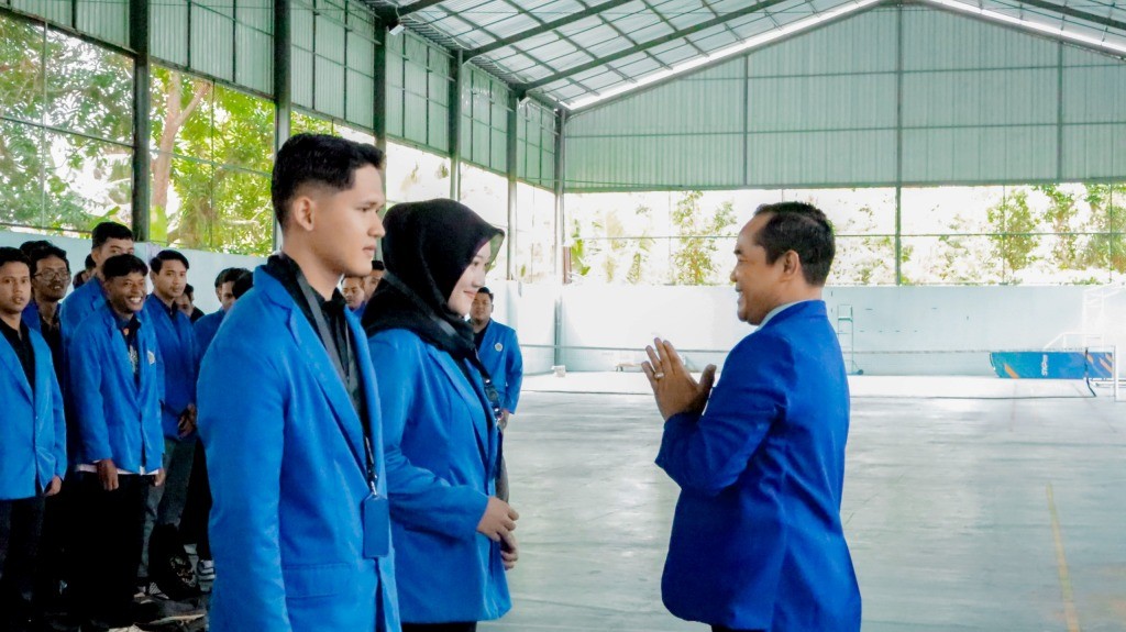 Ketua STKIP PGRI Pacitan, Bakti Sutopo, berjas biru, memberikan salam dengan menangkupkan tangan di depan dada kepada dua perwakilan mahasiswa KKN berjas biru dalam acara pelepasan di dalam gedung aula yang luas. Sejumlah mahasiswa lain berdiri berbaris di latar belakang menyaksikan prosesi tersebut.