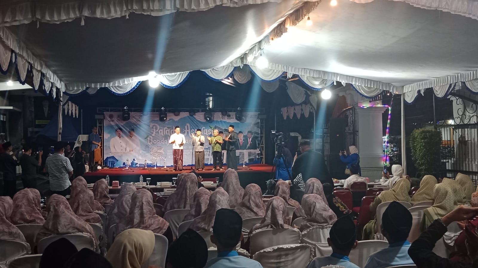 Sebuah panggung di bawah tenda besar yang diterangi lampu sorot, dengan empat orang pria berbaju koko dan bersarung berdiri di atasnya. Di belakang mereka terdapat spanduk besar bertuliskan "PARADE ASPIRASI QOSIDAH REBANA". Di depan panggung, banyak penonton yang duduk di kursi, sebagian besar mengenakan jilbab berwarna cokelat dan peci. Sebuah kamera video di atas tripod merekam acara dari sisi kanan panggung.
