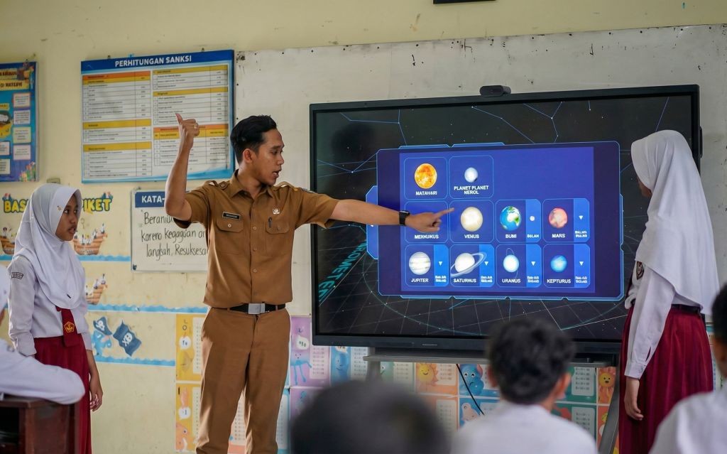 Seorang guru laki-laki, Khrisna Adi Pradana, mengenakan seragam dinas, sedang menunjuk ke layar Interactive Flat Panel (IFP) besar yang menampilkan gambar planet-planet tata surya. Ia sedang mengajar dua siswi berhijab dan siswa lainnya di ruang kelas SDN Arjowinangun, Pacitan.