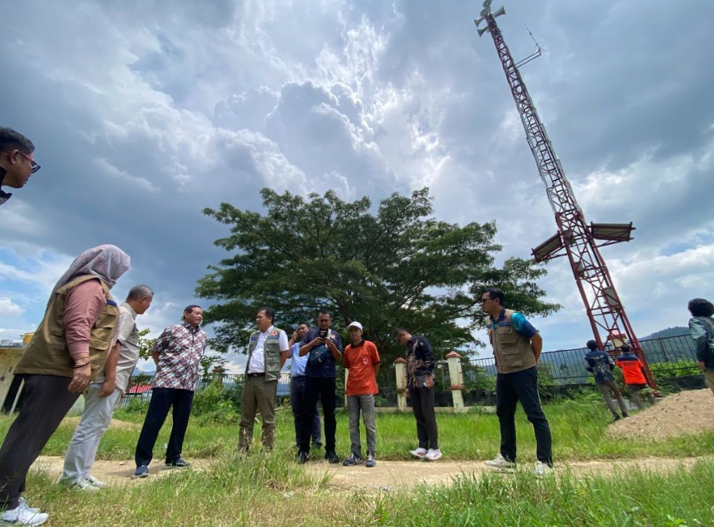 Sebuah foto yang menunjukkan sekelompok pejabat, petugas BNPB, dan warga sedang melakukan pengecekan menara sirene sistem peringatan dini tsunami (EWS) di lapangan terbuka di Desa Kembang, Pacitan, pada Minggu (8/2/2026), dengan latar belakang pohon besar dan langit berawan.