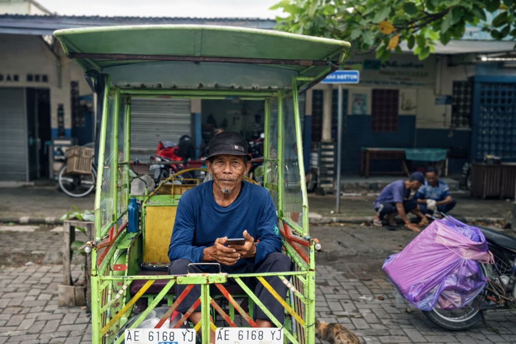 Mujiono, seorang pengemudi becak motor lanjut usia, duduk beristirahat di atas kendaraannya sambil menunggu penumpang di kawasan Pasar Minulyo, Pacitan.