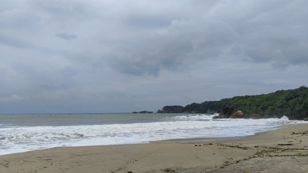 Pemandangan Pantai Soge dari atas jembatan, memperlihatkan hamparan pasir pesisir, aliran muara sungai, dan ombak laut selatan di bawah awan mendung.