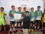 Seorang pejabat pria dari Disbudparpora Pacitan menyerahkan piagam penghargaan "JUARA 1" kepada tim putra pemenang yang mengenakan jersey tosca dan medali di atas podium dalam acara Kejurkab Kabaddi Pacitan 2026, dengan latar belakang spanduk kejuaraan.