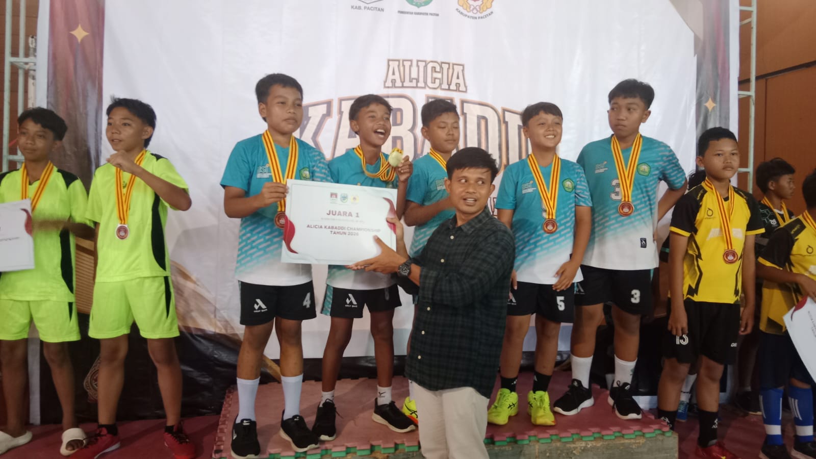 Seorang pejabat pria dari Disbudparpora Pacitan menyerahkan piagam penghargaan "JUARA 1" kepada tim putra pemenang yang mengenakan jersey tosca dan medali di atas podium dalam acara Kejurkab Kabaddi Pacitan 2026, dengan latar belakang spanduk kejuaraan.