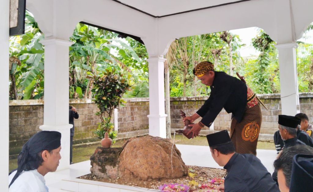 Seorang pria berbusana adat Jawa lengkap (beskap hitam, blangkon, jarik) sedang menuangkan air dari kendi tanah liat ke atas pusara batu besar di dalam sebuah bangunan cungkup makam yang berlantai keramik putih. Beberapa orang lain tampak menyaksikan di latar belakang.
