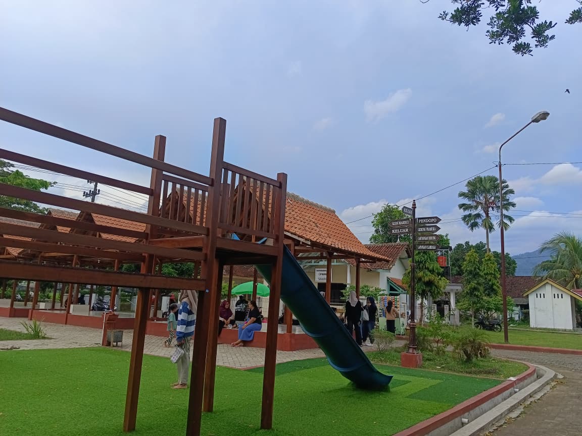 Fasilitas seluncuran berwarna biru yang terpasang pada struktur panjat kayu besar di atas rumput sintetis di area Nol KM Lorok. Terlihat papan penunjuk arah kayu dan beberapa orang duduk di gazebo di latar belakang.