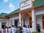 Suasana peresmian Masjid Muniroh Hamad Al-Salim bantuan WAMY di Kelurahan Ploso, Pacitan, dengan warga dan tokoh masyarakat berkumpul di depan pintu masuk bangunan masjid berwarna putih dengan aksen oranye dan atap hijau.