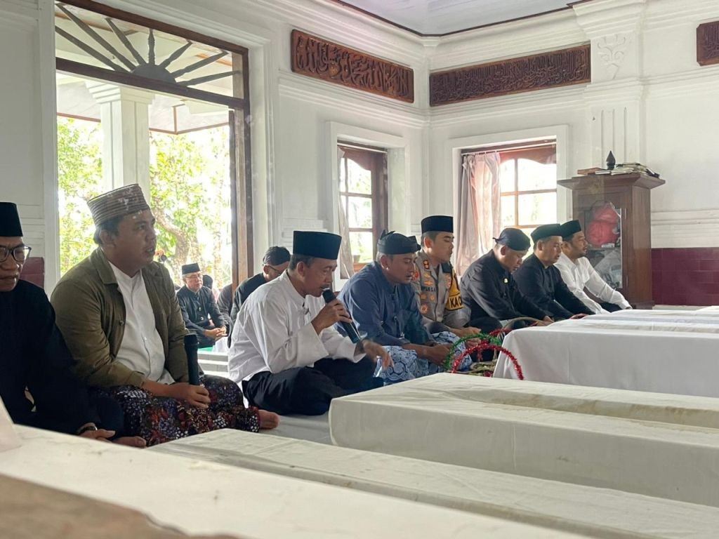 Seorang pria berpeci dan berbaju koko putih memegang mikrofon memimpin doa, duduk di samping makam tertutup kain putih. Ia dikelilingi oleh pejabat lain, termasuk seorang polisi dan pria berpakaian adat Jawa, yang mendengarkan dengan saksama.