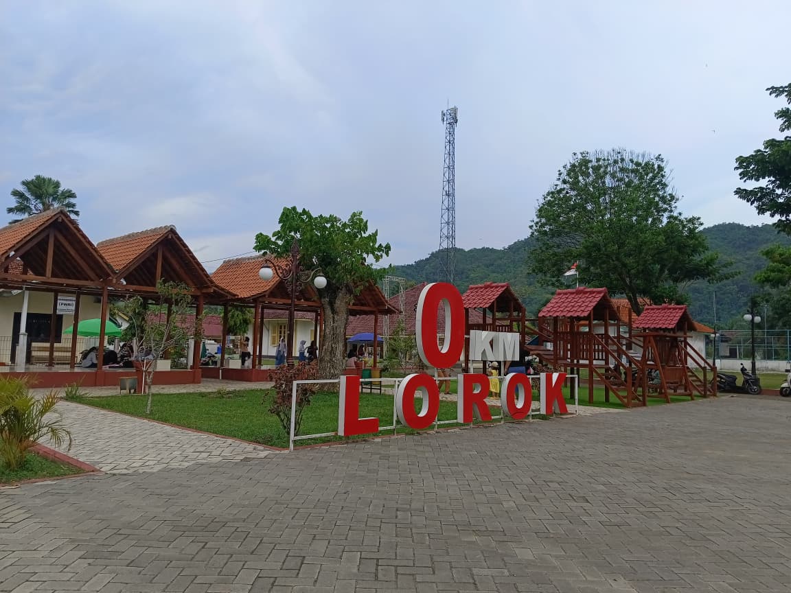 Pemandangan lanskap area rekreasi publik Nol Kilometer Lorok di Pacitan, menampilkan tulisan merah besar "0 KM LOROK", deretan gazebo beratap genteng, dan fasilitas bermain anak kayu berlatar belakang perbukitan dan menara telekomunikasi.
