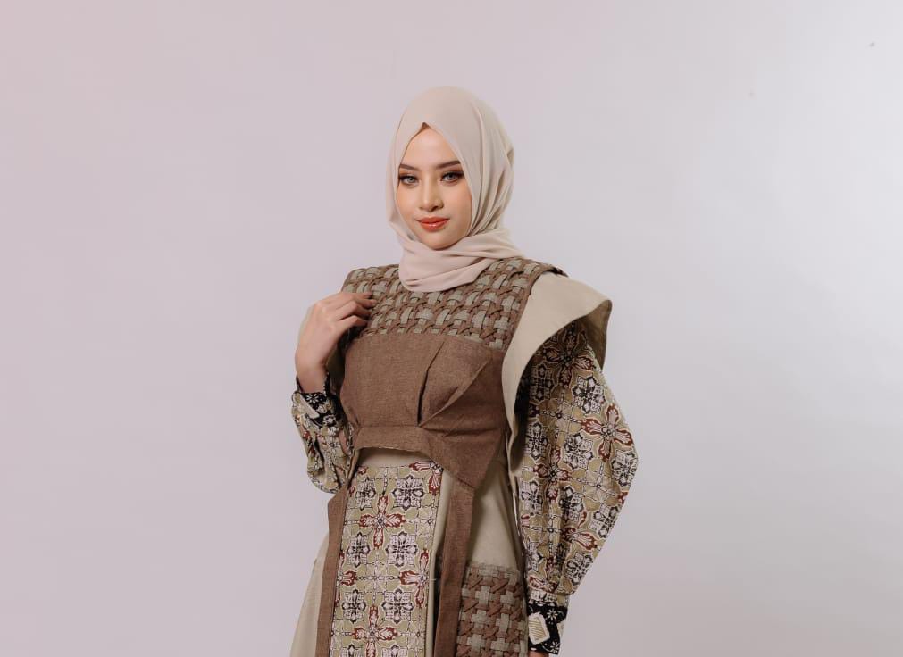 Seorang model wanita berhijab berpose mengenakan busana muslimah modern bernuansa warna bumi (earth tone). Pakaian tersebut merupakan kombinasi tunik dan rompi (vest) yang memadukan kain polos dengan kain bermotif batik atau tenun bertekstur, produk dari Kinana Fashion Pacitan.