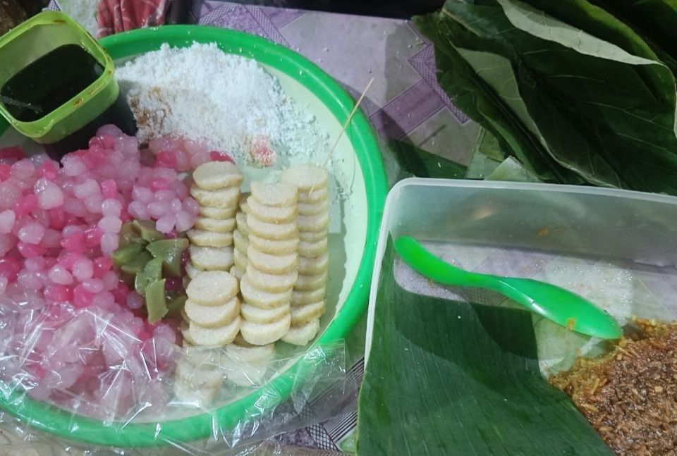 Close-up jajanan cenil merah muda yang mengkilap dengan siraman gula merah cair dan taburan kelapa putih di atas daun pisang.