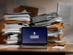 Sebuah pandangan dari dekat di atas meja kantor yang berantakan, di mana sebuah laptop menampilkan layar log-in, hampir sepenuhnya terkepung oleh tumpukan besar dan tidak rapi dari dokumen kertas yang dijepit dan amplop manila. Pemandangan ini menyoroti risiko keamanan data di lingkungan kerja yang padat karya dan berantakan