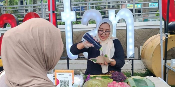 Tangan penjual sedang menaburkan parutan kelapa di atas butiran cenil merah muda yang beralaskan daun pisang