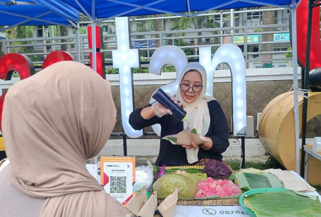 Tangan penjual sedang menaburkan parutan kelapa di atas butiran cenil merah muda yang beralaskan daun pisang
