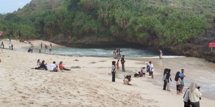 Sejumlah wisatawan berjalan-jalan dan duduk santai di hamparan pasir putih Pantai Srau, Pacitan, dengan latar belakang deburan ombak dan tebing karang pada H+3 libur Lebaran.