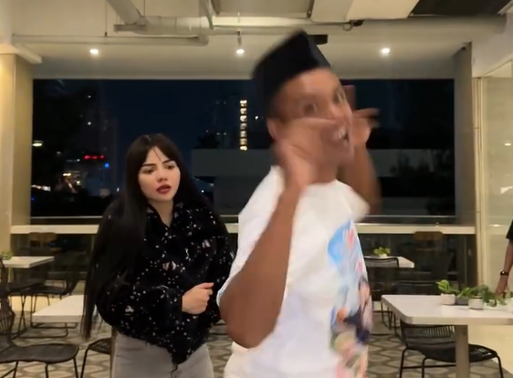Akip Pahala (kanan), kreator konten asal Pacitan mengenakan sarung putih dan peci hitam, tersenyum lebar saat berjoget bersama Dinar Candy (kiri) yang mengenakan kaus putih, di sebuah pujasera pada malam hari.