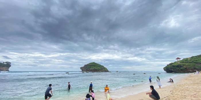 Wisatawan bersantai di Pantai Watu Karung, Pringkuku, Pacitan, pada sore hari, menampilkan tebing karang ikonik dan peselancar.