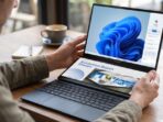 Seorang pria memegang laptop dua layar ASUS Zenbook DUO UX8407AA yang menampilkan Windows Copilot di layar atas dan materi desain di layar bawah di atas meja kayu.