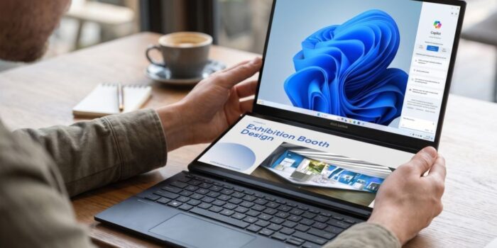 Seorang pria memegang laptop dua layar ASUS Zenbook DUO UX8407AA yang menampilkan Windows Copilot di layar atas dan materi desain di layar bawah di atas meja kayu.