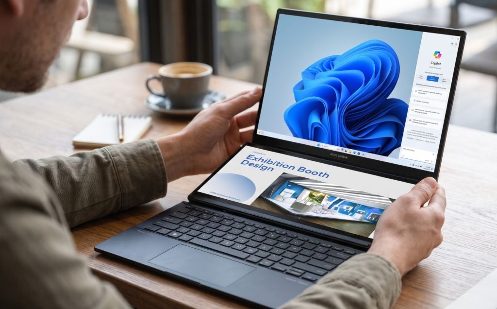 Seorang pria memegang laptop dua layar ASUS Zenbook DUO UX8407AA yang menampilkan Windows Copilot di layar atas dan materi desain di layar bawah di atas meja kayu.