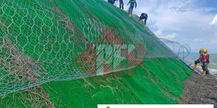 Pekerja konstruksi memasang lembaran geomat dan menancapkan pasak jangkar pada permukaan tanah lereng yang telah diratakan.