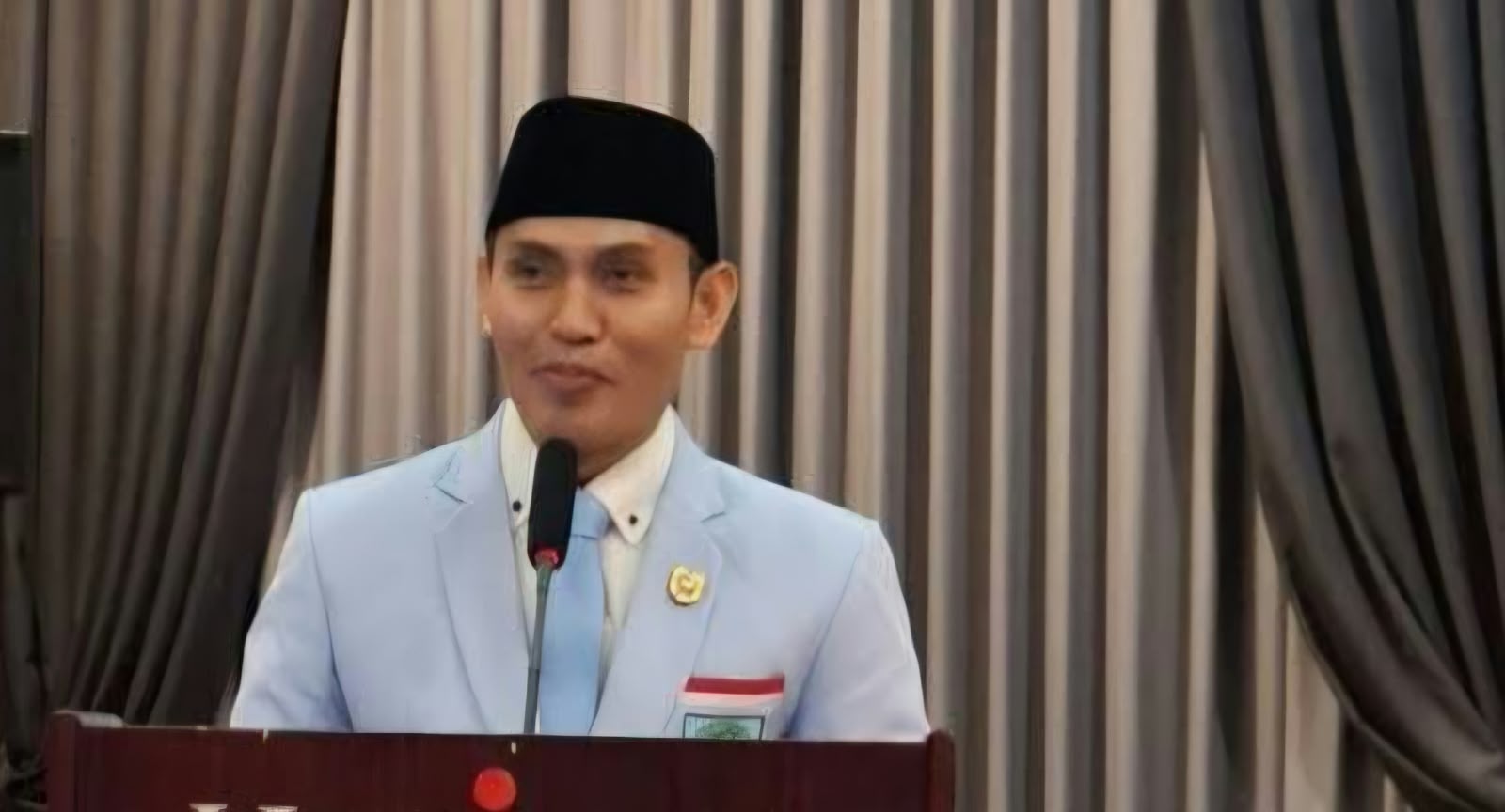 Ketua Umum DPP BKPRMI KH. Nanang Mubarok mengenakan jas biru muda dan peci hitam saat berbicara di depan mikrofon podium.